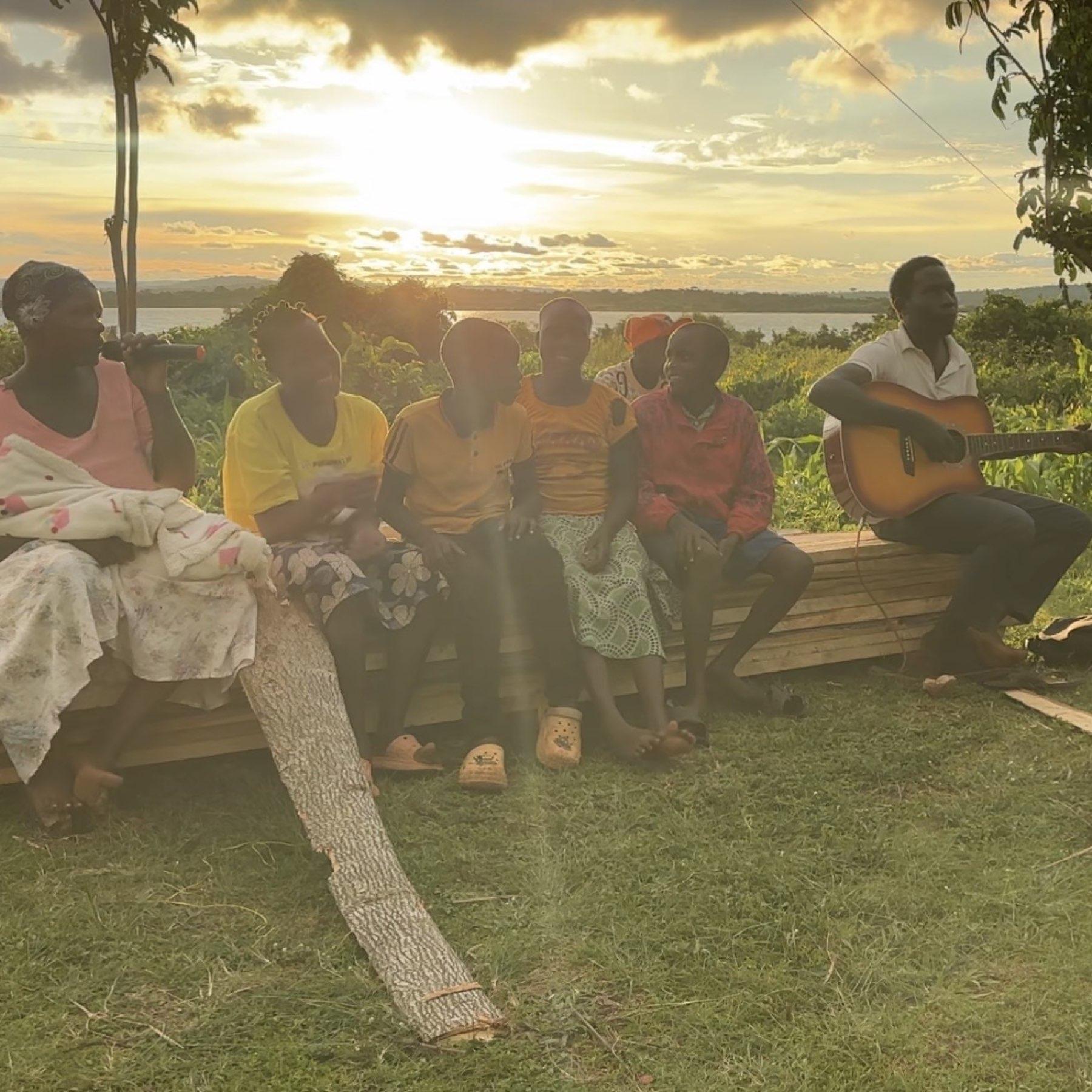 Estate families singing and playing guitar together on a bench at sunset, with Lake Victoria behind them.